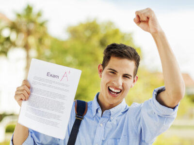 Portrait of happy student clenching fist while holding exam paper with A+ grade on university campus. Horizontal shot.